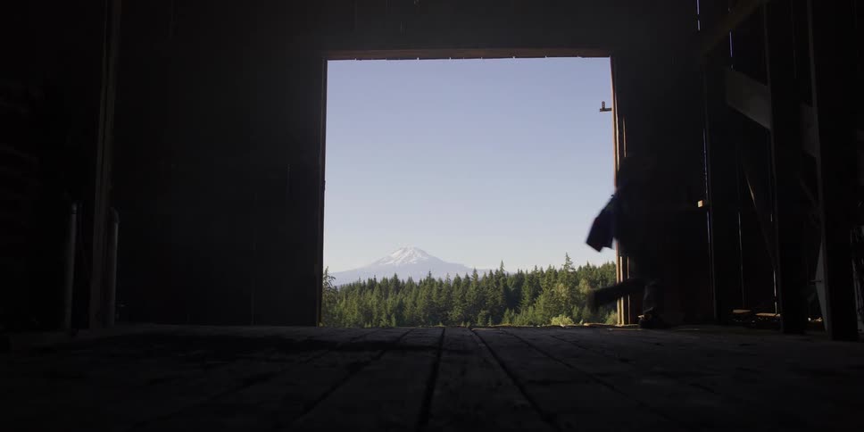 A silhouetted child in a cowboy hat runs across a dark barn floor with a view of a snow-capped mountain outside