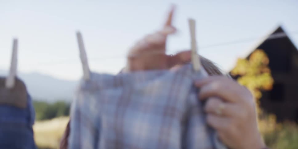 A thoughtful woman hangs a plaid shirt on a clothesline in a sunlit country field in slow motion