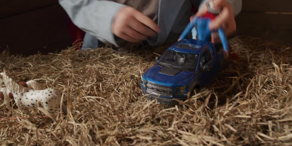 An imaginative young boy wearing a cowboy hat and superhero cape plays with his toy truck and action figures in a hay-filled barn loft