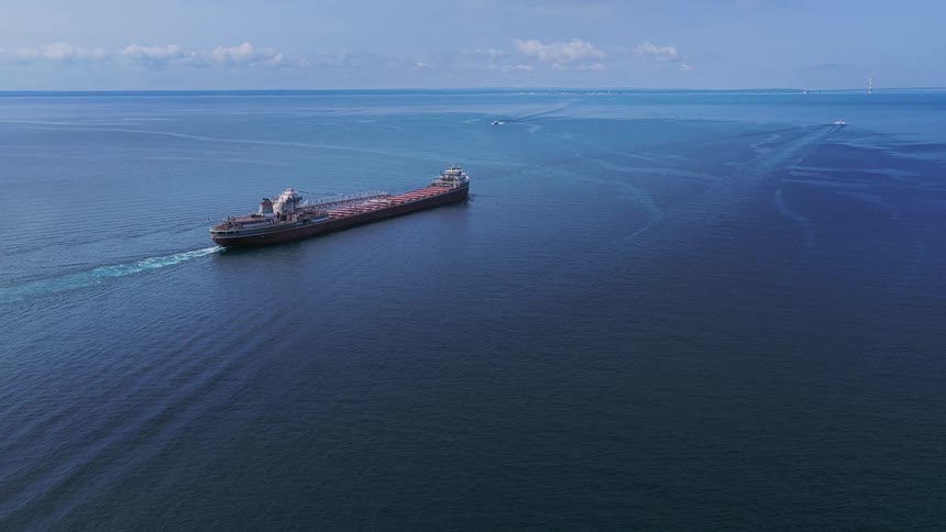 A high-angle aerial shot captures a massive cargo freighter ship sailing across the vast, deep blue ocean on a sunny day