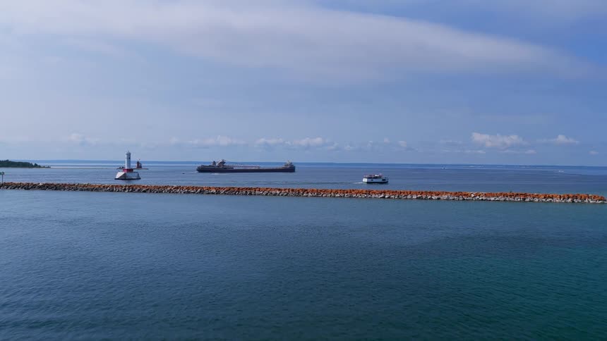An aerial view flies over a calm blue harbor towards a rocky breakwater with a lighthouse, revealing a large freighter on the open water