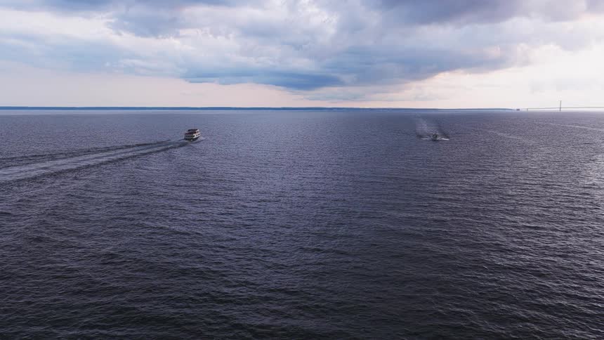 A wide aerial view shows a tour boat and another vessel sailing across a vast, dark ocean under a dramatic, cloudy sky