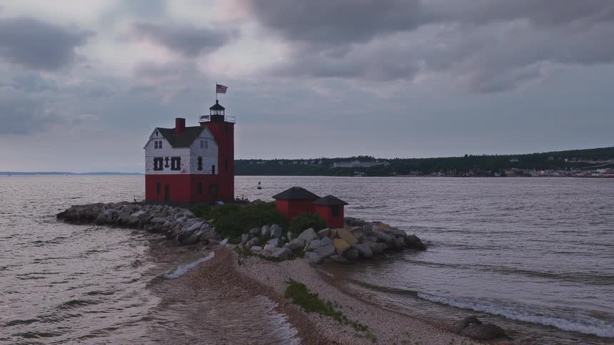 The iconic red and white Round Island Lighthouse stands on a rocky point as gentle waves lap the shore under a cloudy evening sky