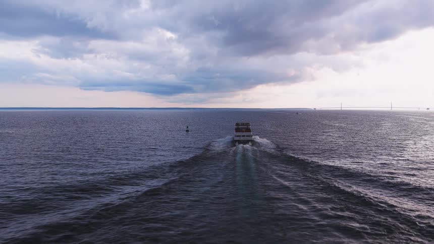 Cinematic aerial view of a ferry sailing across the sea towards a distant bridge under a dramatic, overcast sky