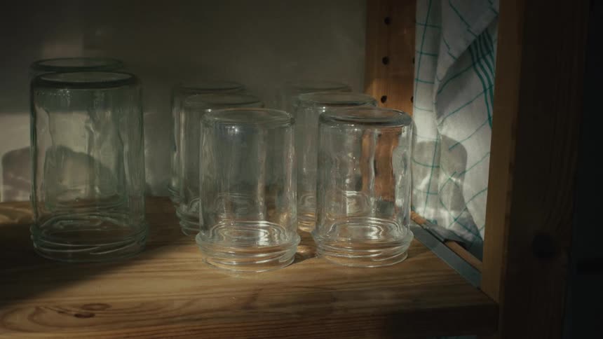 Sunlight streams into a kitchen, casting moving shadows across clean, empty glass jars resting on a wooden shelf