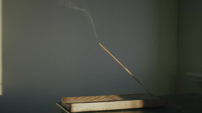 A stick of incense burns in a wooden holder, releasing a gentle stream of smoke in the sunlight