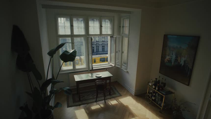 A cinematic, high-angle view from inside a sunlit apartment shows a yellow tram passing by on the city street below through an open window