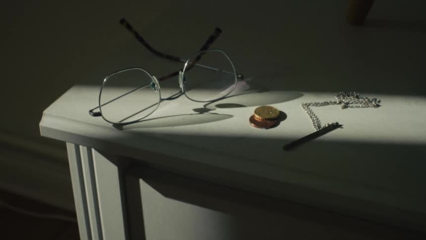 A moody still life of personal items, including glasses and a necklace, resting on a white dresser in a stark beam of sunlight