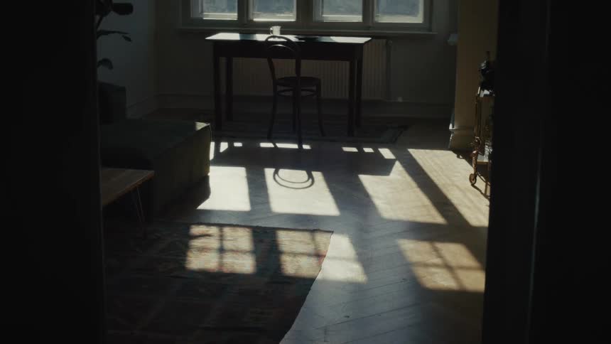 A slow tilt up from a dark hallway reveals a quiet room bathed in sunlight, with shadows from the window stretching across the wooden floor