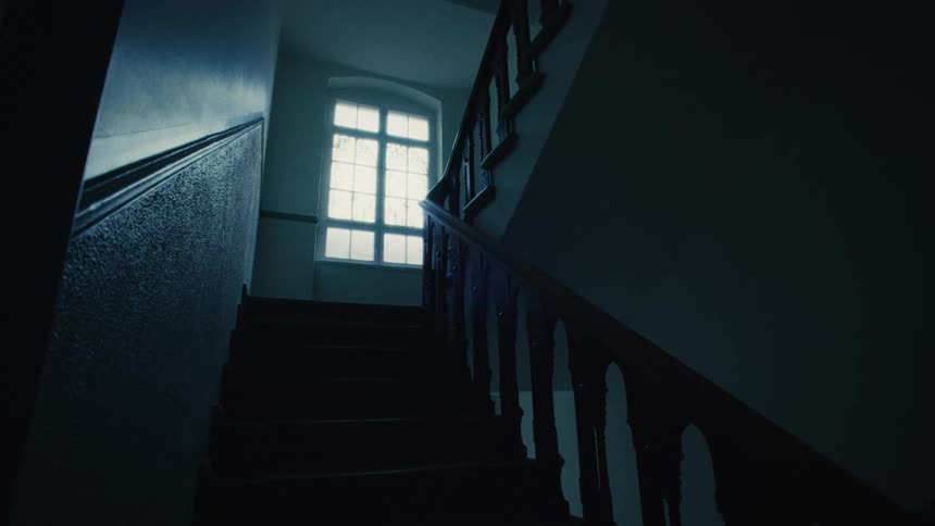 A low angle shot of a creepy, dark staircase in an old building, with light shining through the window at the top