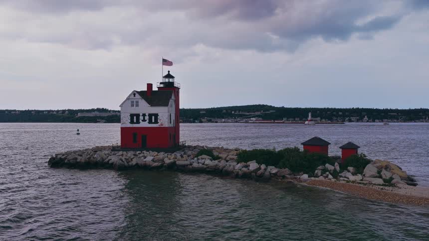 The iconic red and white Round Island Lighthouse stands on a rocky islet in the Straits of Mackinac under a cloudy sky