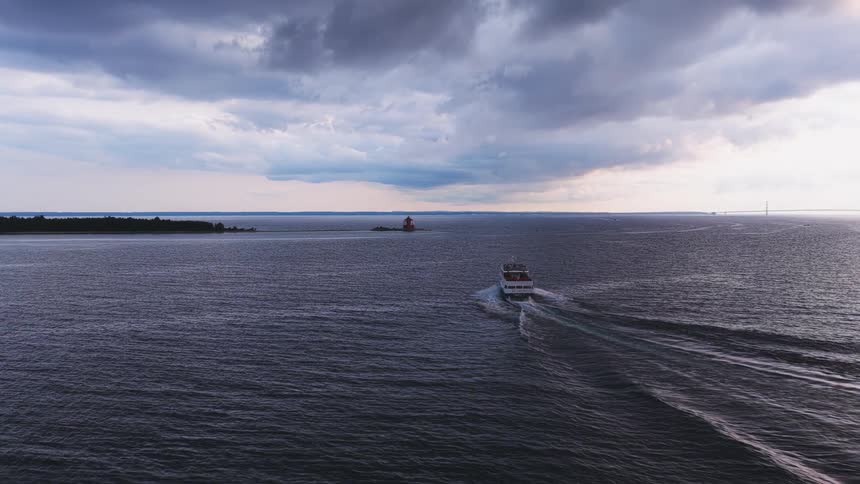 An aerial view follows a tour boat sailing across a wide expanse of water towards a lighthouse under a dramatic, cloudy sky