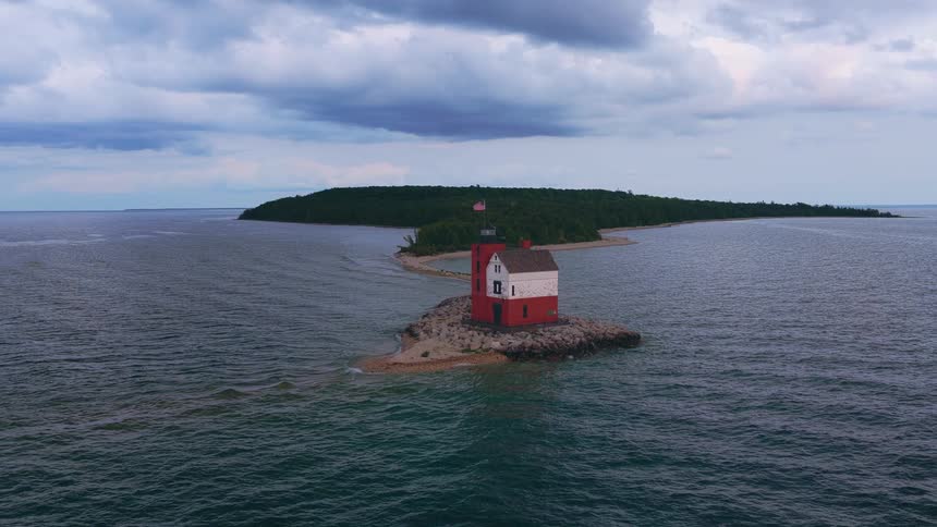 A stunning aerial view of a red and white lighthouse on a small rocky islet connected by a sandbar to a forested island under a cloudy sky