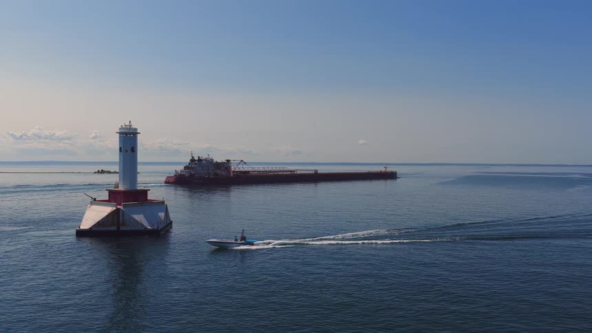 A small boat cruises across the calm blue water past a lighthouse, with a large cargo freighter in the background on a sunny day