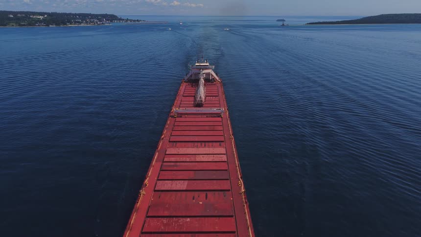An aerial view follows a massive red cargo ship sailing across a vast blue lake on a clear, sunny day