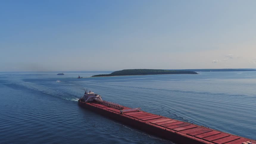 An aerial view of a large red cargo freighter sailing on the calm blue water towards a distant island