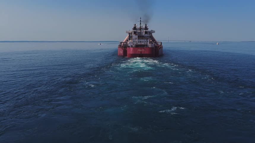 A large cargo ship sails across the open water, leaving a trail of smoke and a churning wake behind it under a clear blue sky