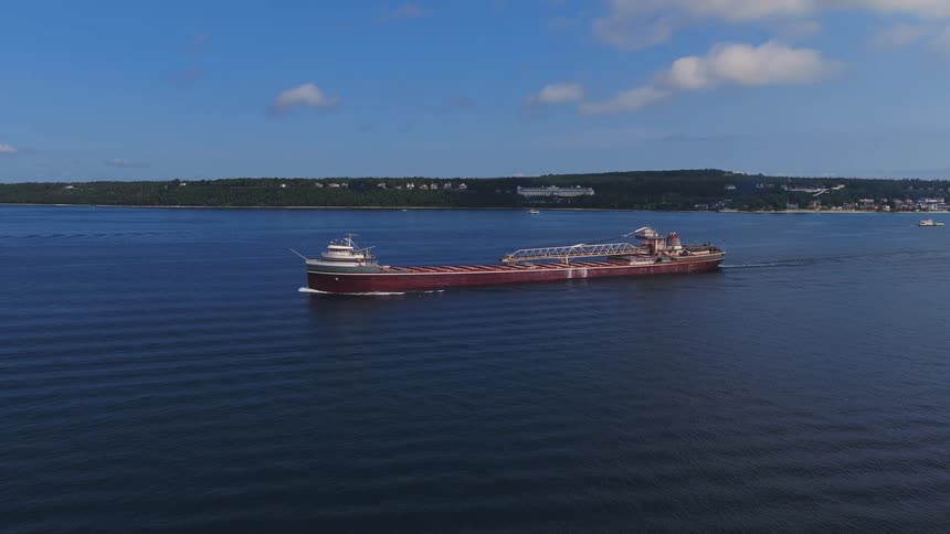 A majestic Great Lakes freighter glides across the serene blue waters near a lush, green coastline under a partly cloudy sky
