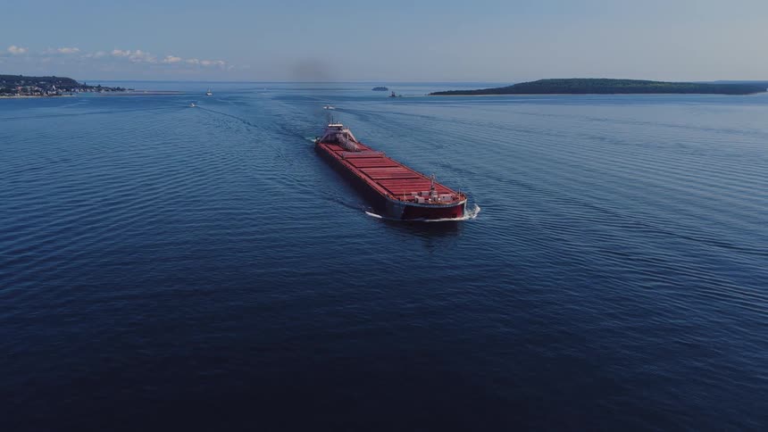 An aerial view follows a massive red cargo freighter sailing across a calm blue lake on a clear day