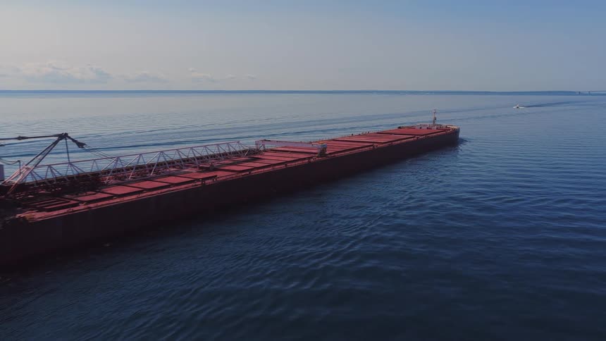 Aerial footage tracks a massive red cargo ship sailing smoothly through the calm, open blue waters on a clear day
