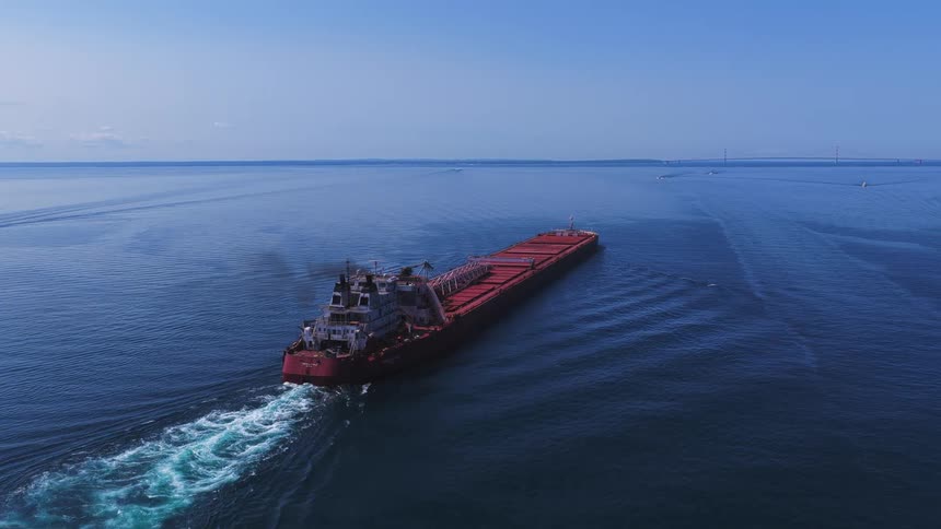 An aerial view follows a massive red cargo freighter sailing across the calm, deep blue water on a clear sunny day towards a distant bridge