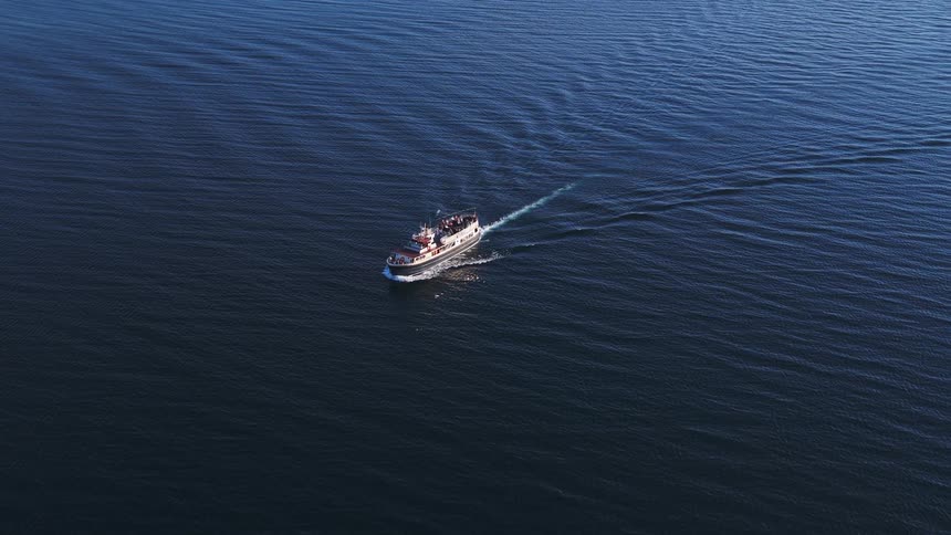 An aerial view of a tour boat sailing across the deep blue sea, leaving a gentle wake on the calm water