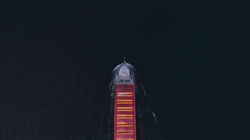 A vibrant red and yellow cargo freighter glides through the dark blue sea in a striking top-down aerial shot