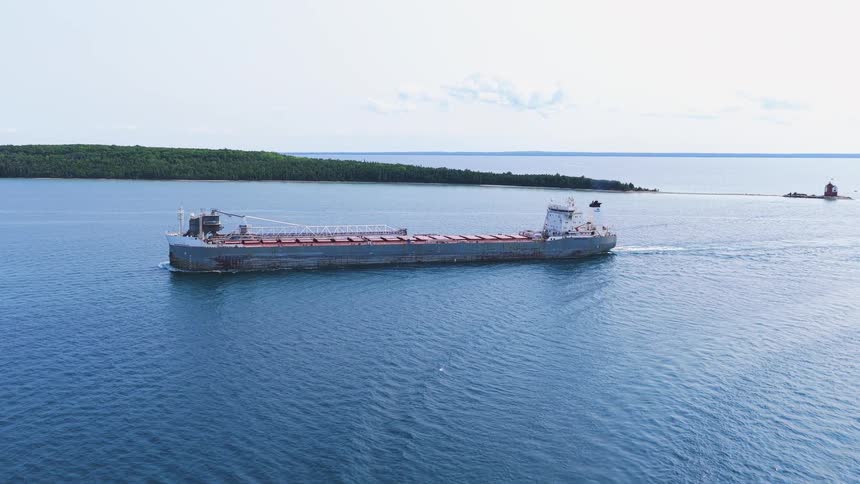 An aerial view captures a massive cargo freighter sailing on the calm blue waters of the Great Lakes, accompanied by smaller tour boats near a forested island