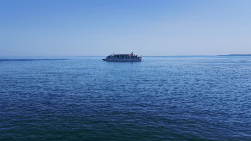 An aerial view slowly approaches a large cruise ship sailing on the vast, calm blue ocean under a clear sky