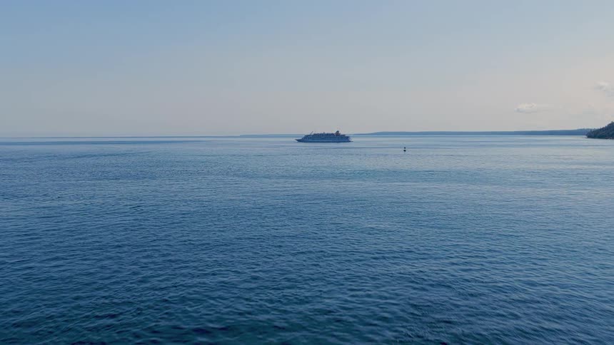 Aerial footage glides past a solitary lighthouse towards a cruise ship sailing on the vast, calm blue ocean under a clear sky