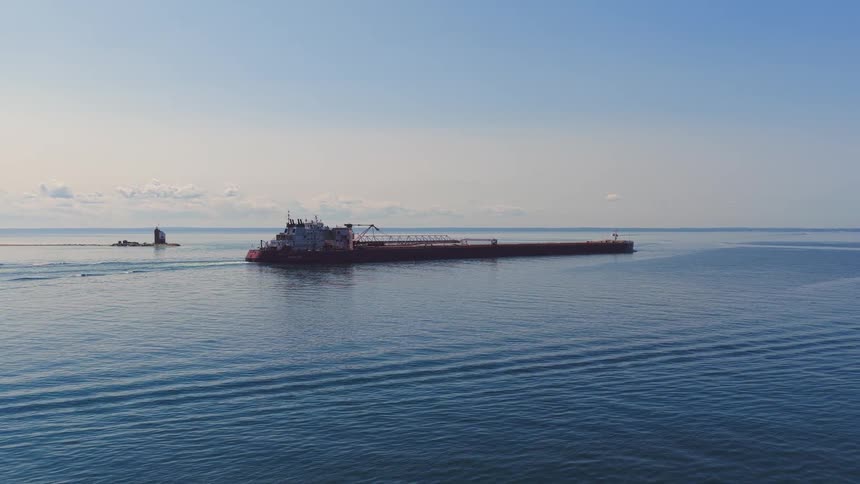 Aerial view of a large industrial barge and tugboat combination sailing across a calm blue lake past lighthouses on a sunny day