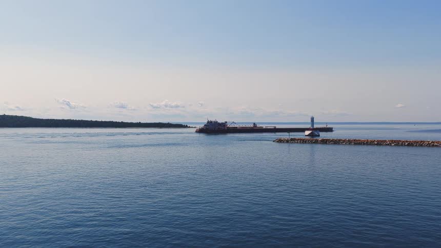 Aerial dolly shot flying over sailboats towards a large cargo ship passing a lighthouse and jetty on a clear, sunny day