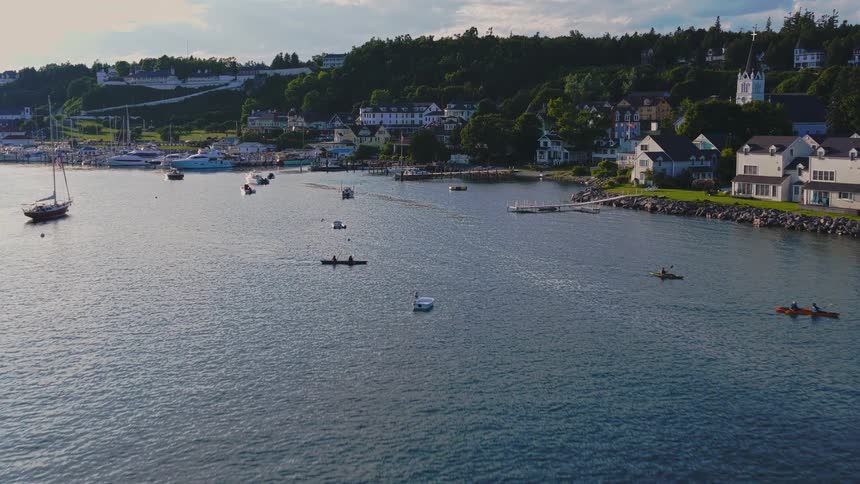 An aerial view flying over a picturesque harbor town with sailboats, yachts, and kayakers on the water during a beautiful summer evening