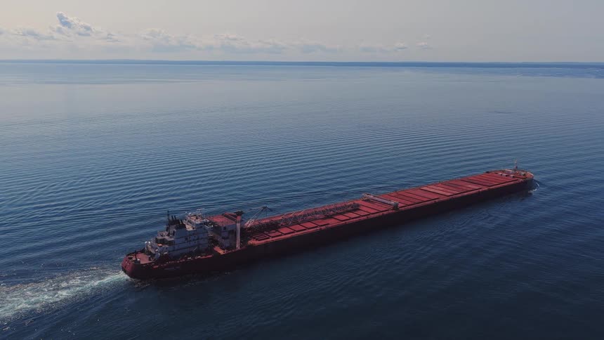 Aerial reveal of a large red cargo freighter sailing across a vast, calm blue lake on a sunny day