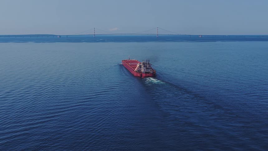 An aerial view shows a massive red freighter sailing across the deep blue waters of the Great Lakes with the majestic Mackinac Bridge in the distant background
