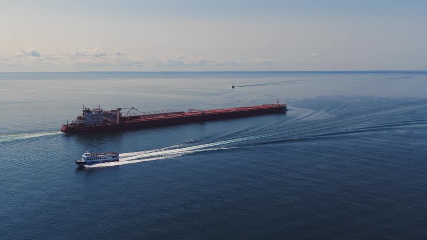 A smaller white passenger ferry speeds past a massive red cargo freighter as they both travel across the vast, calm blue ocean