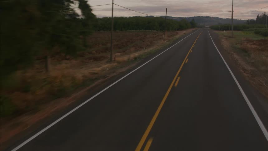 An aerial view following an empty two-lane country road surrounded by trees and fields at dusk