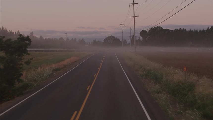 An aerial view slowly glides down an empty, misty country road surrounded by foggy fields at dawn