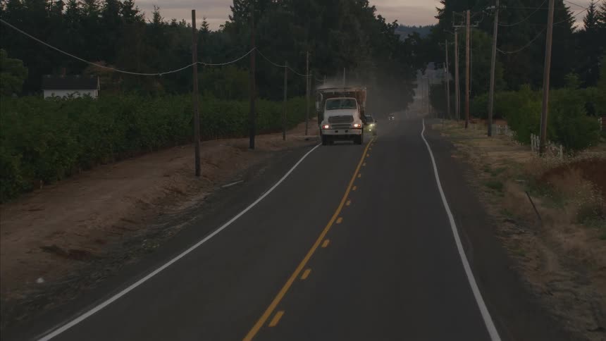 An aerial view shows a large dump truck followed by an SUV driving towards the camera on a rural country road at dusk
