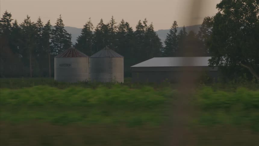 A tranquil tracking shot passes a rural farm with two grain silos and a barn at twilight, with evergreen trees and mountains in the background