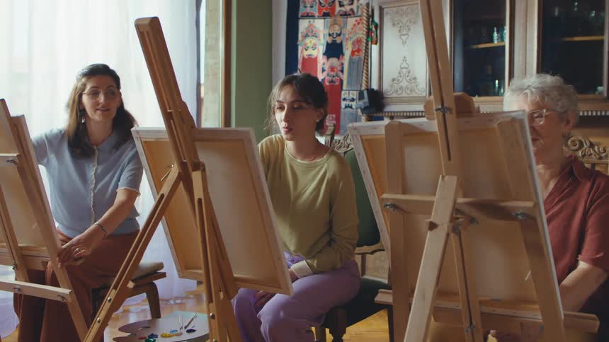A multi-generational group of women listens to their instructor during a painting class in a cozy art studio