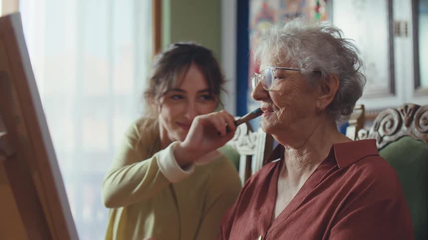 A granddaughter playfully dabs her grandmother's cheek with a paintbrush while they enjoy painting together in a sunlit room