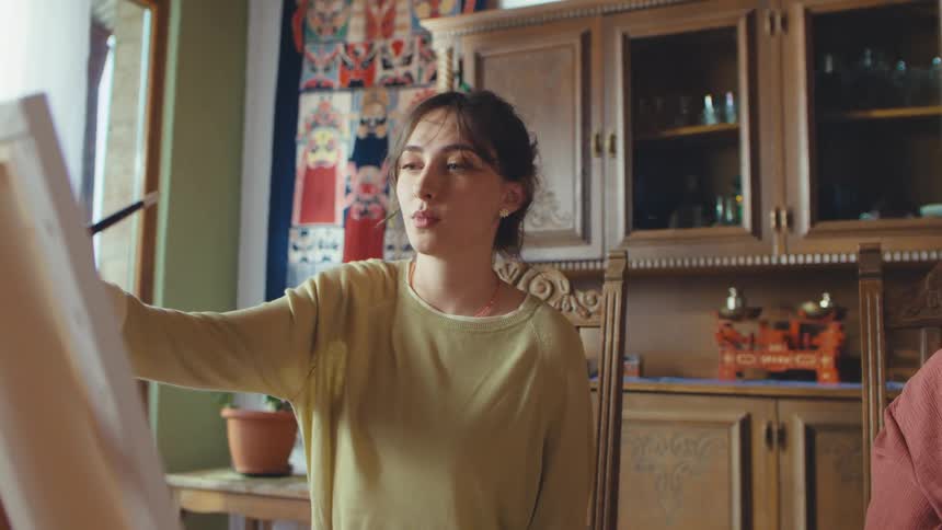 An art teacher guides a young student with her painting as the camera pans to an elderly woman happily creating her own artwork in a cozy home studio