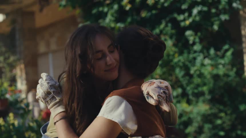 A young woman in overalls shares a loving hug with an older woman in a beautiful sunlit garden