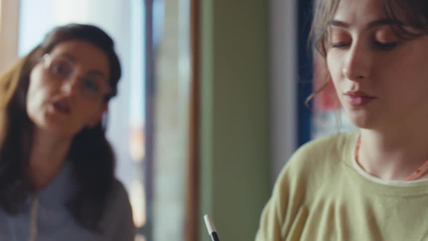 A young artist carefully applies paint to a canvas while her teacher offers guidance in a sunlit art studio