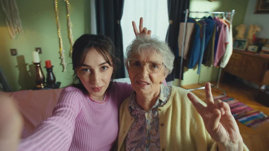 A granddaughter and her grandmother take a playful selfie video, smiling and making peace signs at the camera while sitting on a couch