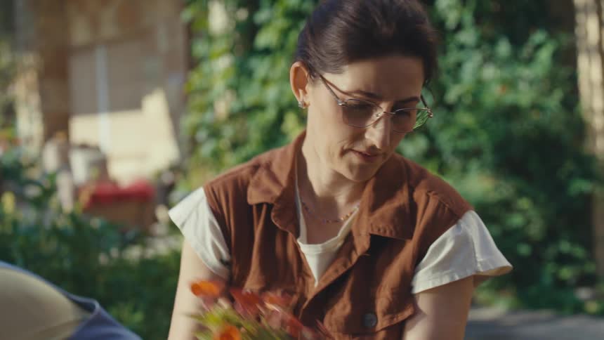 A woman with glasses and gardening gloves holds a red flower plant before potting it in her sunlit garden