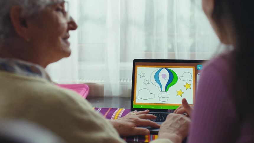 A cheerful grandmother and her granddaughter enjoy a digital coloring activity together on a laptop at home