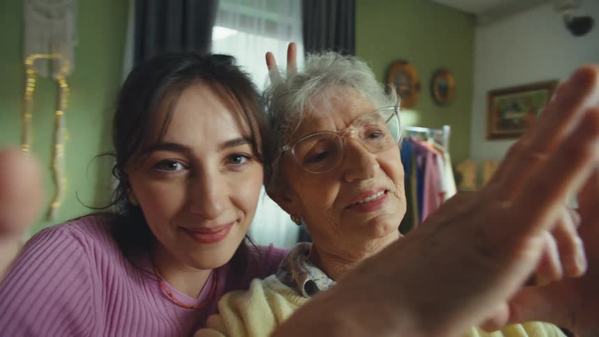 A loving granddaughter and her cheerful grandmother take a selfie, making playful gestures like bunny ears and heart shapes for the camera