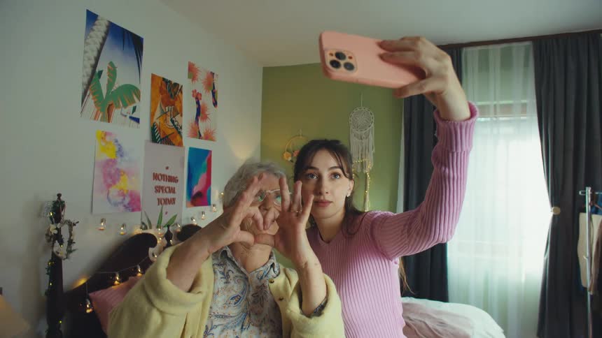A cheerful granddaughter and her grandmother take a selfie together in a decorated bedroom, making a heart shape with their hands for the camera